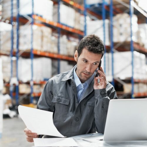 A young man in a warehouse taking orders on his cellphone and recording them on his laptop