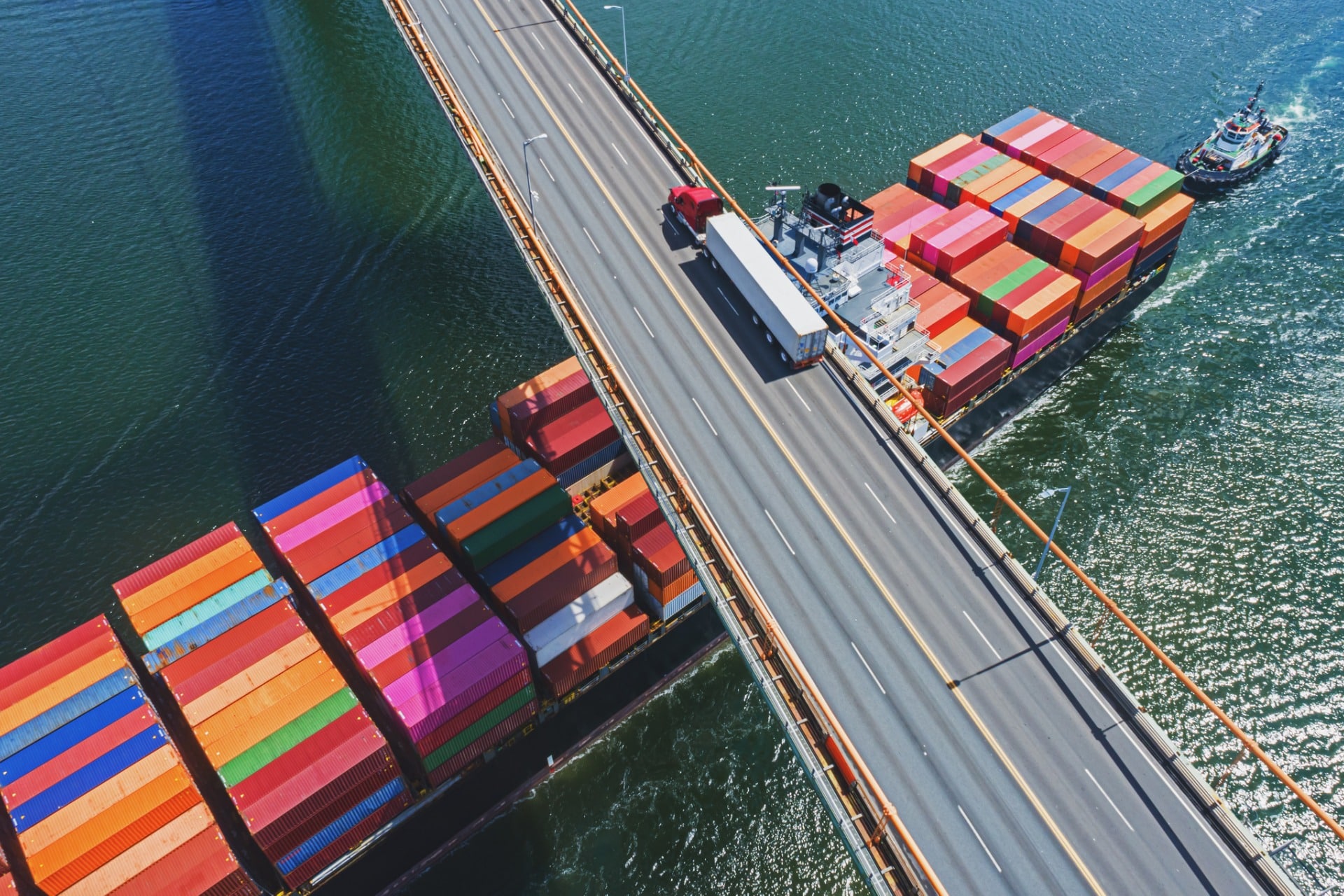 This is a photo of a ship covered in multi-colored shipping containers passing underneath a bridge that has a semi-truck driving across it. This photo represents the supply chain and recent difficulties managing the supply chain.