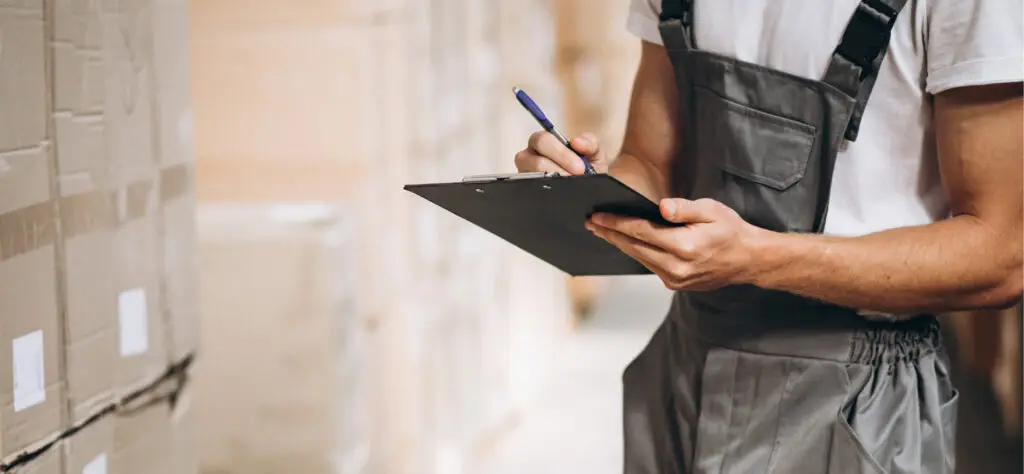 A person in gray overalls holds a clipboard and pen, checking or writing on paper while standing next to stacked cardboard boxes in a logistics warehouse.